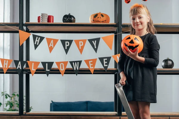 adorable kid with pumpkin standing on ladder at hanging flags with happy halloween inscription at home