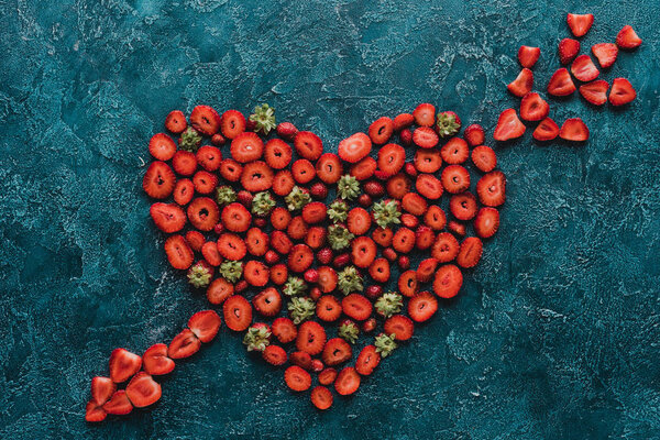 top view of heart pierced with arrow sign made of strawberries on blue concrete surface