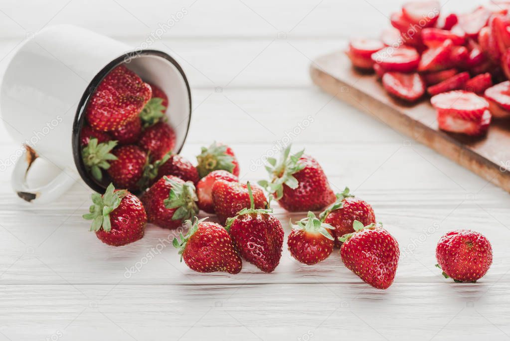 ripe strawberries spilled from mug onto white wooden surface with cutting board