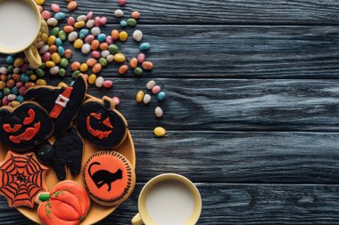top view of plate with halloween cookies, colorful candies, cups with milk on wooden table  clipart