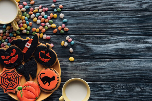 top view of plate with halloween cookies, colorful candies, cups with milk on wooden table 