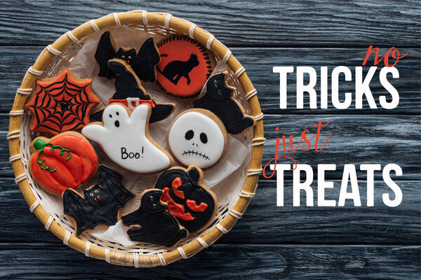 top view of wicker basket with homemade spooky halloween cookies on wooden table  with "no tricks just treats" lettering