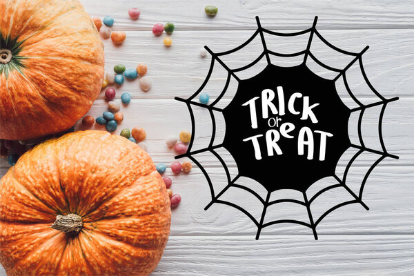 вид сверху на тыквы и красочные конфеты на деревянном фоне с паутиной и надписью "трюк или угощение"
