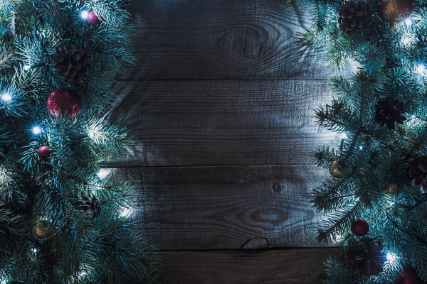 top view of beautiful christmas tree branches with baubles, pine cones and illuminated garland on wooden background 