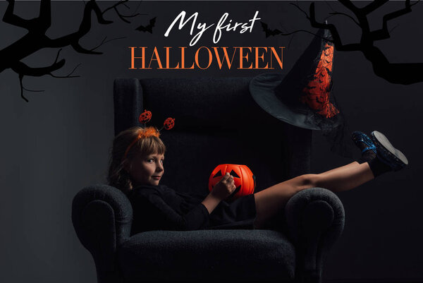 adorable kid in halloween costume resting in armchair in dark room with "my first halloween" lettering 