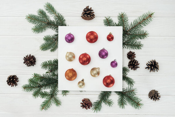 top view of shiny colorful christmas balls on white card, fir twigs and pine cones on wooden surface