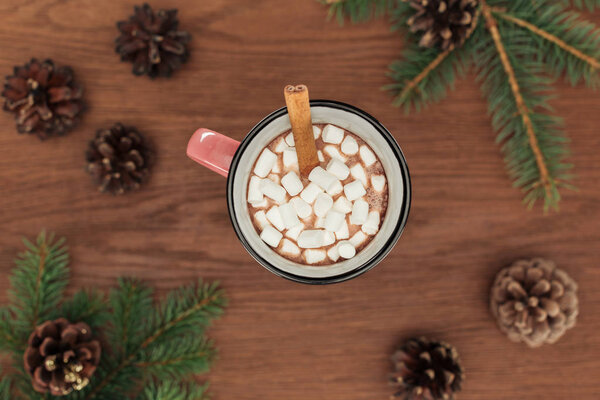 top view of cup with hot chocolate, marshmallows, cinnamon stick, coniferous twigs and pine cones on wooden table