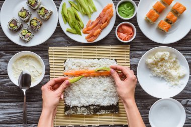 cropped shot of person cooking delicious sushi roll with salmon, rice, cucumber, avocado and nori  clipart