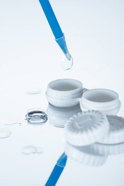 close up view of container, contact lenses and tweezers on white backdrop with reflection