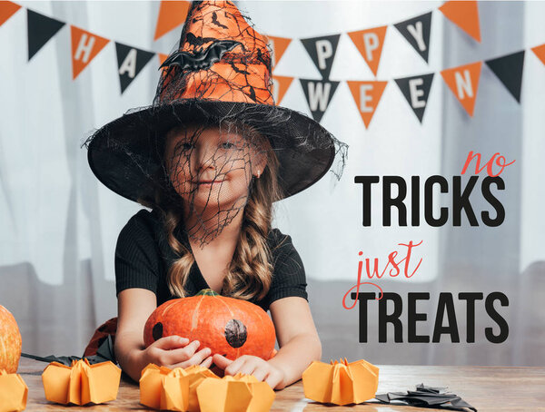 portrait of adorable kid in witch halloween costume at tabletop with pumpkins at home with "no tricks just treats" lettering