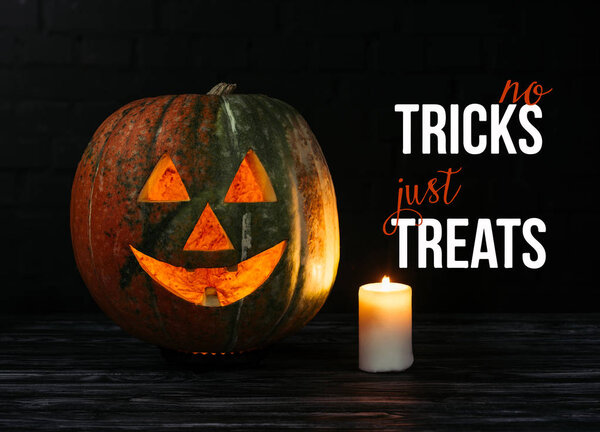 spooky halloween carved pumpkin with candle on wooden table on black background with "no tricks just treats" halloween lettering