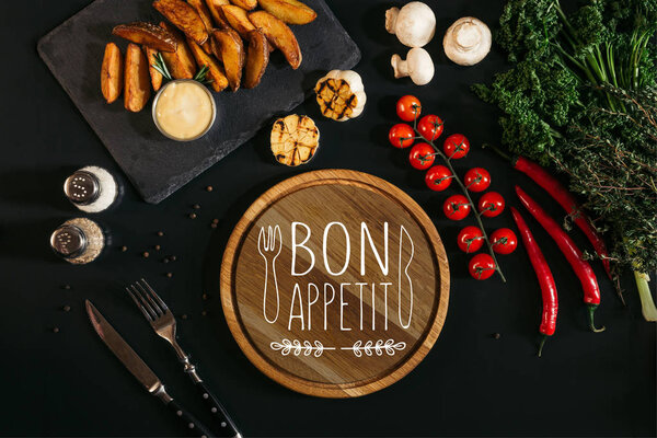 top view of wooden board  with "bon appetit" lettering, vegetables and baked potatoes on black 