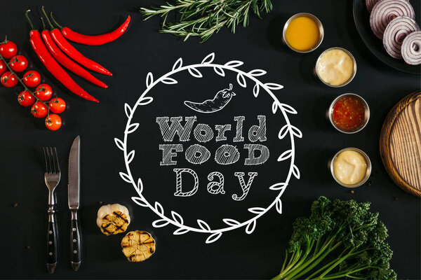 вид сверху на различные соусы, жареный чеснок, вилку с ножом и свежие овощи с травами на черном фоне с надписью "Всемирный день еды"
