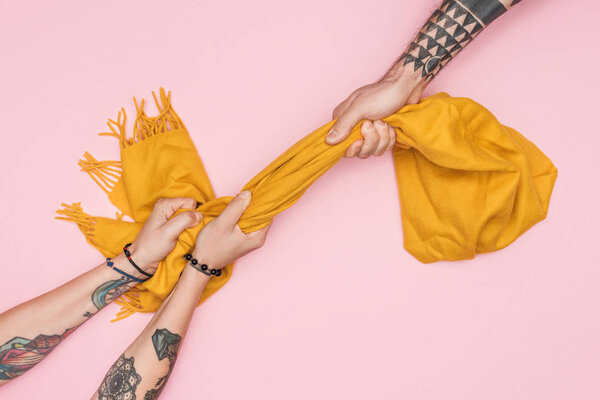 частичный вид татуированных шопоголиков тянущих желтый шарф изолированный на розовый
