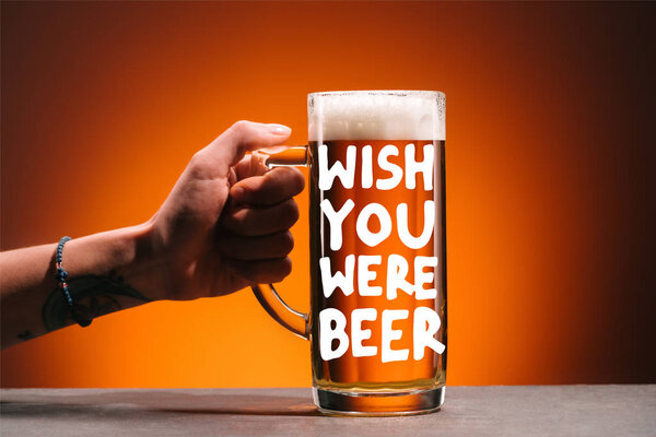 cropped shot of woman holding glass of cold beer with foam on orange backdrop with "wish you were beer" inspiration