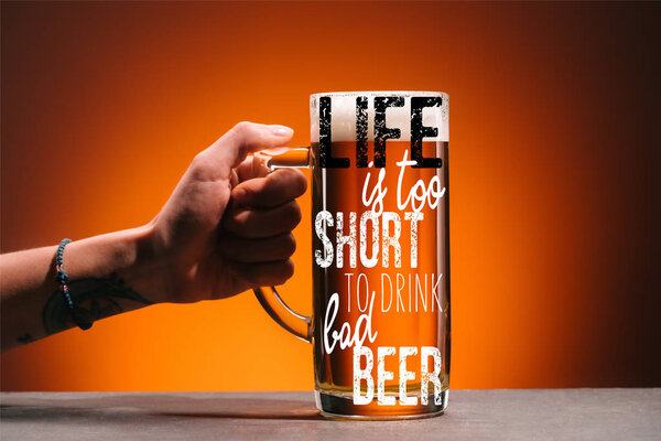 cropped shot of woman holding glass of cold beer with foam on orange backdrop with "life is too short to drink cheap beer" inspiration