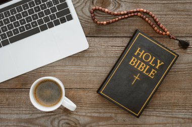 top view of holy bible with beads, laptop and coffee on wooden table clipart