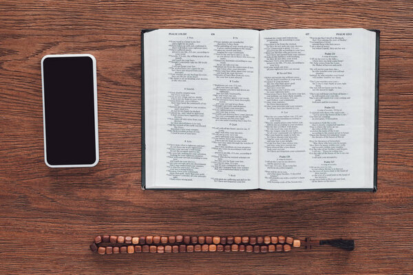 top view of opened holy bible with beads and smartphone on wooden table