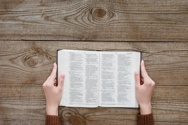 cropped shot of woman holding opened holy bible on wooden surface