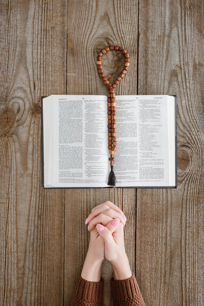 cropped shot of woman praying with holy bible and beads on wooden table