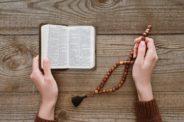 cropped shot of woman with holy bible holding beads on wooden surface