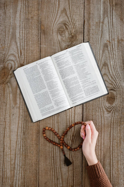 cropped shot of woman with holy bible holding beads on wooden table