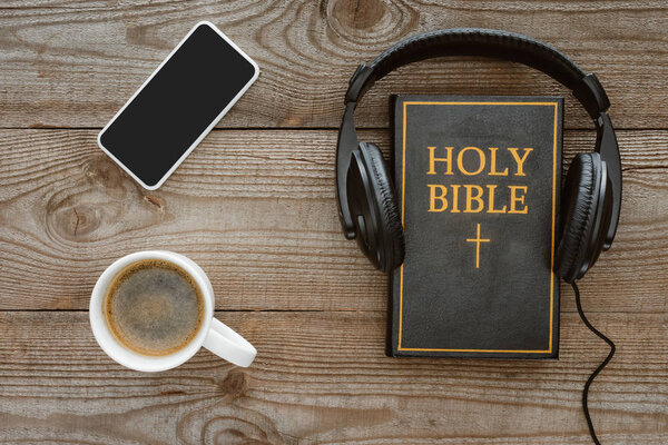 верхний вид Священной Библии с наушниками, смартфоном и кофе на деревянном столе
