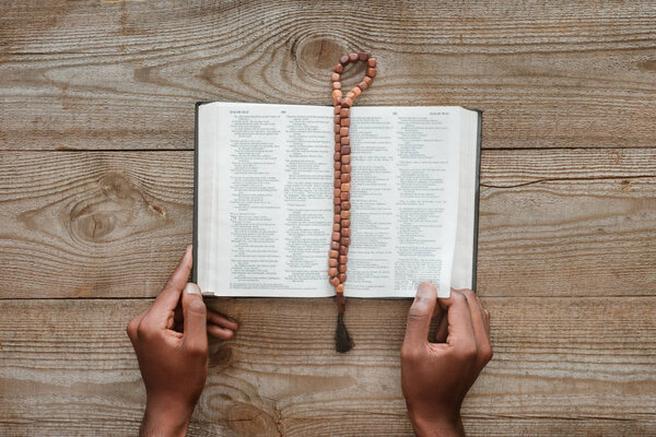 cropped shot of african american man reading holy bible with beads lying above on wooden table