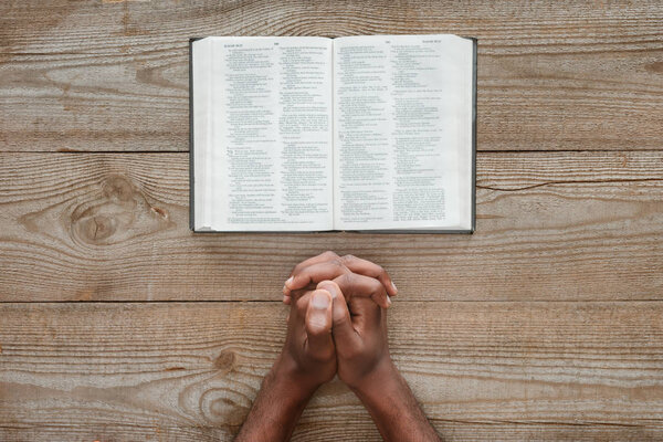 cropped shot of african american man praying with holy bible on rustic wooden table