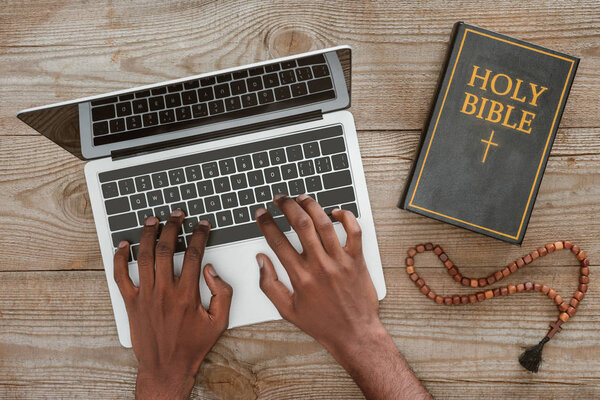 cropped shot of man working with laptop with holy bible and beads on wooden tabletop