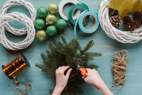 частичный вид женщины, делающей рождественский венок ручной работы на голубом деревянном столе
