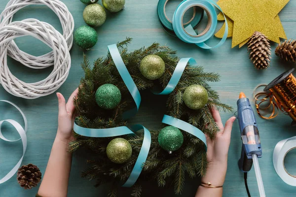 在蓝色木表面上用丝带和玩具装饰的手工圣诞花环的妇女短款拍摄 — 图库照片