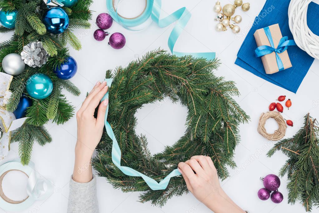 cropped shot of woman decorating handmade christmas wreath with blue ribbon isolated on white