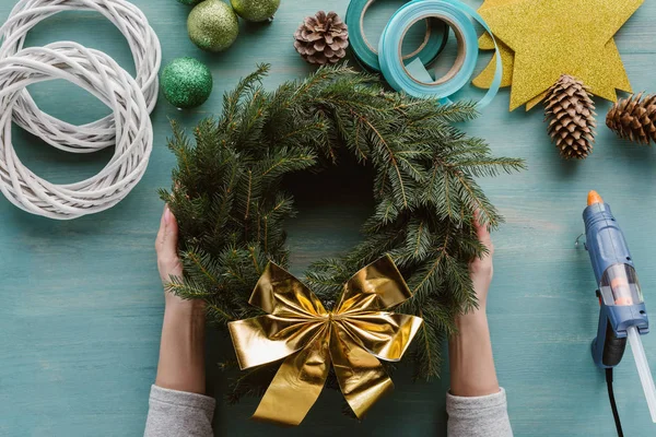 Cropped shot of woman holding handmade pine tree wreath with golden bow and decorations on blue wooden tabletop — Stock Image Cropped Shot Woman Holding Handmade Pine Tree Wreath Golden Bow Royalty Free Stock Photos