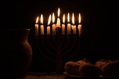 close up view of traditional sweet doughnuts, clay jug and menorah with candles on black background, hannukah holiday concept clipart