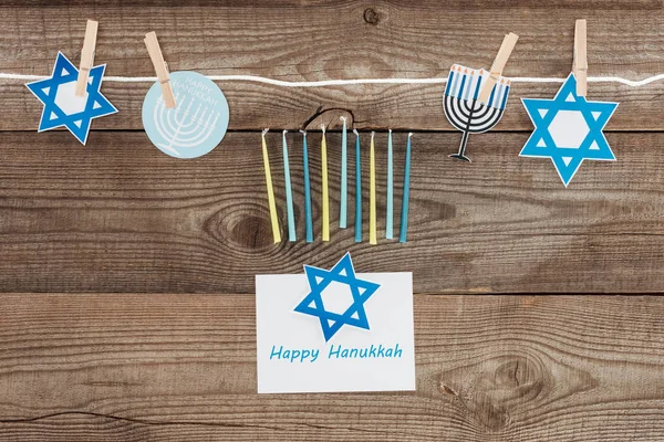 top view of happy hannukah card, candles and holiday paper signs pegged on rope on wooden tabletop, hannukah concept