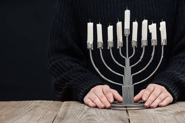 cropped shot of woman standing at wooden tabletop with menorah and candles on black background, hannukah celebration concept
