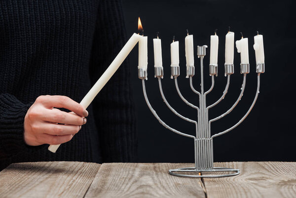 частичный вид женщины, зажигая свечи на меноре на деревянном столе на черном фоне, Hannukah праздничной концепции
