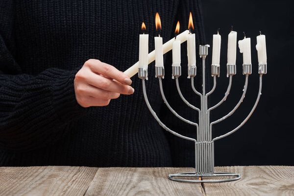 partial view of woman lighting candles on menorah on wooden tabletop on black backdrop, hannukah holiday concept