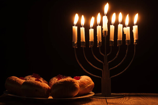 close up view of traditional sweet donghnuts and menorah with candles on black background, hannukah holiday concept

