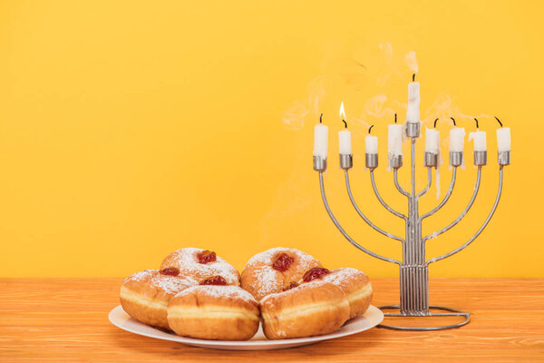 close up view of sweet doughnuts and menorah with candles on wooden surface isolated on yellow, hannukah concept