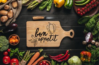 top view of cutting board with knife and organic fresh vegetables around on wooden tabletop, bon appetit lettering clipart