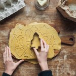 Partial view of woman cutting raw dough with cookie cutter while preparing christmas cookies on wooden surface