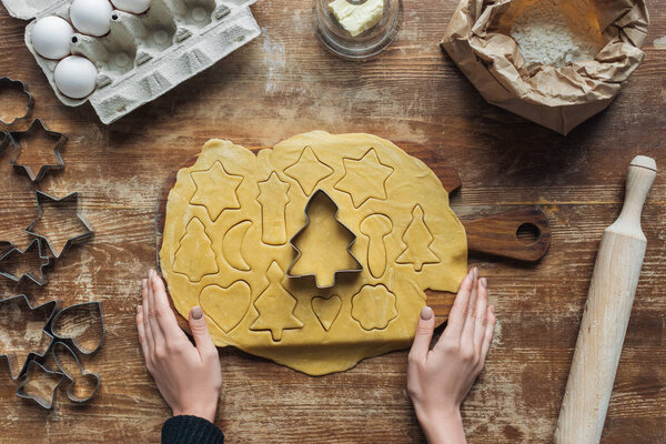 cropped shot of female hands, raw dough, ingredients for christmas cookies bakery and cookie cutters on wooden tabletop