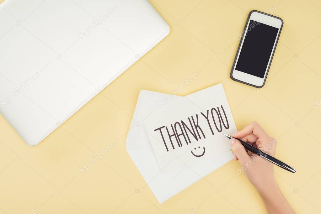 cropped person holding pen above white postcard with thank you lettering, smartphone and laptop isolated on yellow background