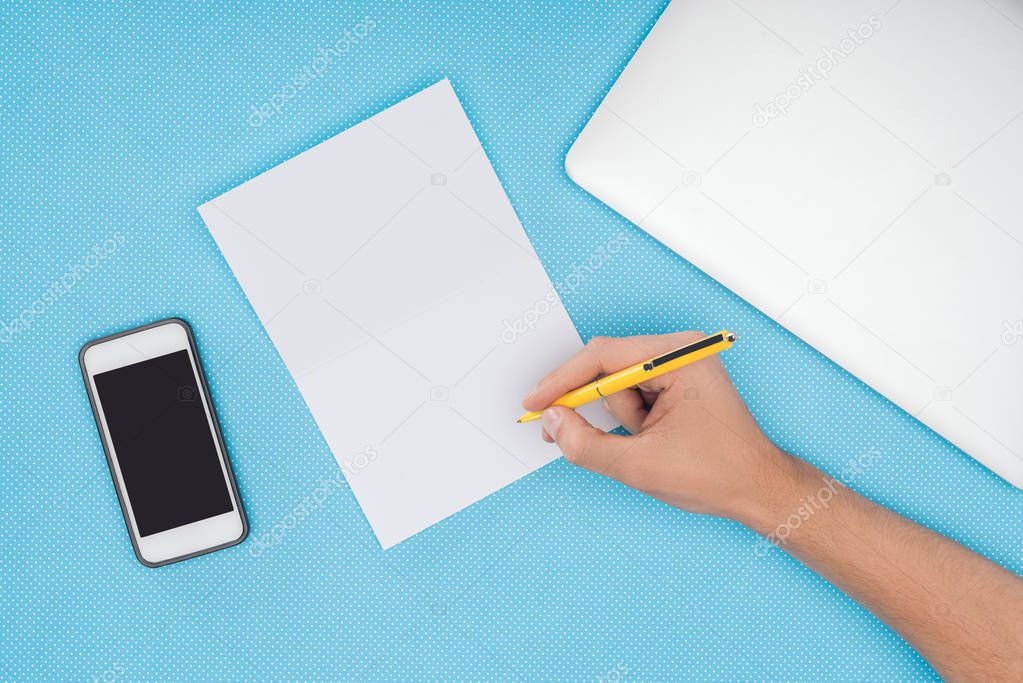 cropped view of hand over blank notebook with smartphone and laptop isolated on blue background