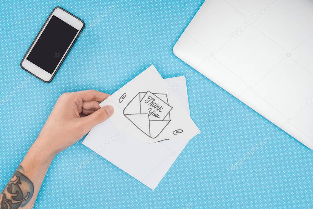 cropped person holding white card with thank you lettering sticking out of hand drawn envelope and smartphone isolated on blue background