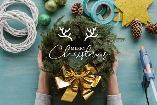 Cropped shot of woman holding handmade pine tree wreath with golden bow and decorations on blue wooden tabletop with "merry christmas" lettering — Stock Image Cropped Shot Woman Holding Handmade Pine Tree Wreath Golden Bow Stock Picture
