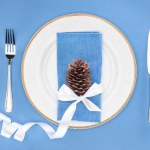 Verhoogde weergave van plaat met dennenappel omwikkeld door feestelijk lint en vork met mes geïsoleerd op blauw