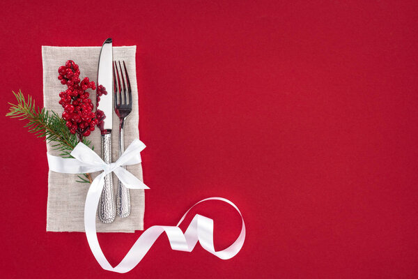 elevated view of plate with pine branch, berries, fork and knife wrapped by ribbon isolated on red